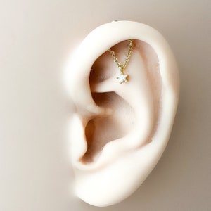 May include: A gold-colored chain earring with a small, white star pendant. The earring is designed to drape across the ear, creating a delicate and stylish look. The star pendant adds a touch of whimsy to the jewelry.