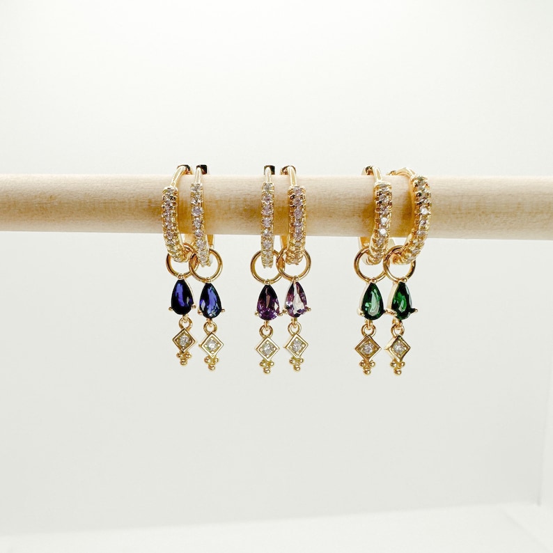 May include: Three pairs of gold hoop earrings with dangling charms. Each pair features a different colored gemstone: purple, green, and blue. The hoops are adorned with small clear stones.