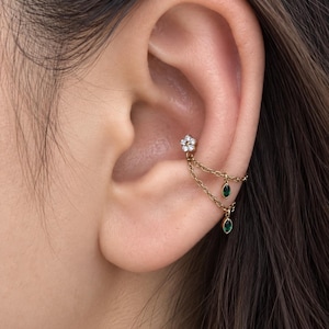 Puede incluir: Un pendiente tipo ear cuff de oro que presenta un pequeño perno en forma de flor con piedras transparentes, conectado por dos delicadas cadenas de oro a dos dijes colgantes de piedra verde con corte marquesa en el cartílago de la oreja.