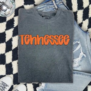 Puede incluir: Una camiseta de manga corta gris carbón con la palabra Tennessee en letras grandes de estilo burbuja de color naranja con un patrón de cuadros negros. La camiseta tiene cuello redondo y un aspecto lavado vintage, colocada sobre una superficie.
