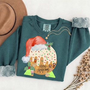 May include: A dark teal long-sleeve sweatshirt featuring a graphic of a Christmas pudding wearing a Santa hat. The pudding is decorated with white icing, red and green sprinkles, and holly. The text Merry Christmas is printed in black script.