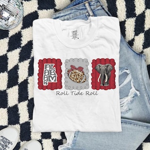 May include: A white t-shirt featuring three rectangular frames with scalloped edges. The left frame contains the word ALABAMA in gray, the center frame shows a leopard print football with a red bow, and the right frame displays a gray elephant.