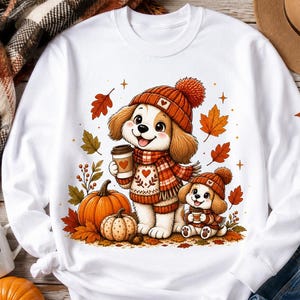 Puede incluir: Una sudadera blanca de manga larga con un gráfico de dos cachorros de dibujos animados con gorros y bufandas de punto naranja, sosteniendo tazas de café. El diseño incluye calabazas naranjas y hojas de otoño dispersas alrededor de los perros.