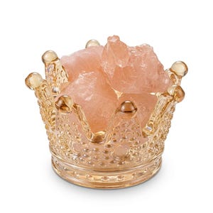 Puede incluir: Un cuenco de cristal con forma de corona, superficie texturizada con cuentas y tono dorado, lleno de varios trozos de cristal de cuarzo rosa en bruto, translúcidos y de color rosa pálido. El recipiente tiene ocho puntas redondeadas.