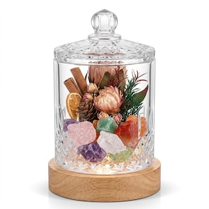 Op de afbeelding: Een doorzichtige glazen pot met een ruitpatroon en bijpassend deksel, rustend op een ronde, lichtgekleurde houten voet. Binnenin bevinden zich diverse ruwe kristallen, gedroogde bloemen, kaneelstokjes, een dennenappel en een gedroogde sinaasappelschijf.