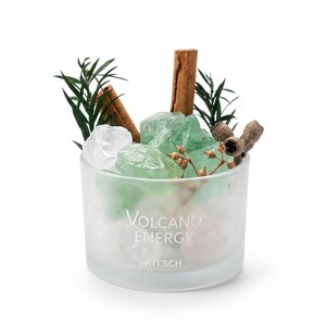 Può includere: Un contenitore in vetro smerigliato con l'etichetta VOLCANO ENERGY KITSCH, riempito con cristalli verdi e trasparenti, due bastoncini di cannella marroni, rametti verde scuro e un piccolo ramo con baccelli marroni essiccati.