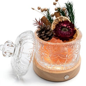 Puede incluir: Un frasco de vidrio transparente con un patrón decorativo en relieve y una tapa a juego, sobre una base circular de madera con luz integrada. El frasco contiene cristales de color naranja, una piña, una rodaja de naranja seca y una flor roja.