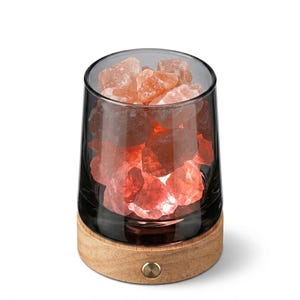 May include: A cylindrical lamp featuring a clear glass vessel filled with raw, orange-pink Himalayan salt chunks. The glass sits on a circular light-coloured wooden base with a small metallic power button centred on the front of the wood.
