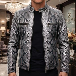 May include: A silver and grey jacket featuring a detailed snakeskin pattern throughout. The jacket has a mandarin collar with a single snap button, a full-length front zip, and two horizontal zipped chest pockets. It is worn over a black shirt.