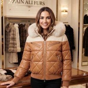 May include: A tan and cream quilted leather jacket featuring a hood with a thick fur trim, a full-length front zipper, two zippered chest pockets, two lower zippered pockets, and ribbed knit cuffs and hem, displayed in a retail store.