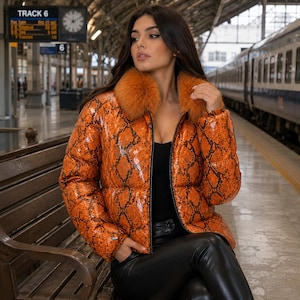 May include: A puffer jacket featuring a vibrant orange and black snakeskin pattern with a thick, fluffy orange faux fur collar. The jacket has a glossy finish, long sleeves, and a front zipper closure, worn over a black top and black trousers.
