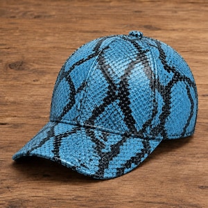 Puede incluir: Una gorra de béisbol con un patrón de piel de serpiente en azul vibrante y negro. La gorra tiene una corona estructurada de seis paneles, una visera curva y un pequeño botón en la parte superior central. El material muestra una textura de escamas.