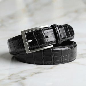 Op de afbeelding: Een zwarte riem met een glanzende krokodil-reliëftextuur en een rechthoekige zilverkleurige metalen gesp. De riem is voorzien van een bijpassende lederen lus en een reeks voorgeponste gaten langs de band voor een verstelbare sluiting.