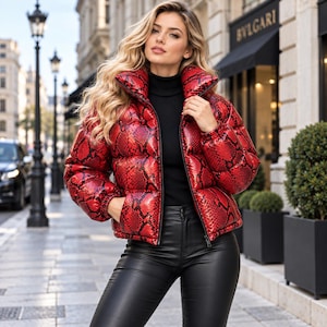 Può includere: Un piumino rosso lucido con motivo a pelle di serpente nera, collo alto e chiusura frontale con zip. La giacca è indossata sopra un maglione a collo alto nero e pantaloni neri, creando un look audace e strutturato dalla finitura vivace.