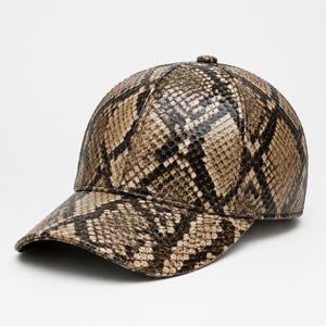 Puede incluir: Una gorra de béisbol con un patrón detallado de piel de serpiente en tonos bronceado, marrón y negro. La gorra tiene una corona estructurada, una visera curva y un pequeño botón en la parte superior central, con una textura de escamas realista.
