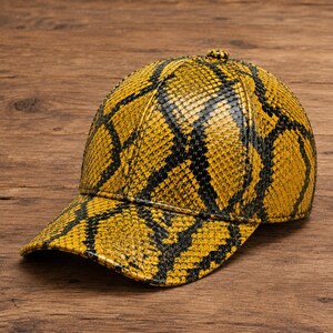 Puede incluir: Una gorra de béisbol con un patrón de piel de serpiente en amarillo y negro vibrante. La gorra tiene una corona estructurada, una visera curva y un pequeño botón en la parte superior central. El material texturizado imita escamas de reptil.