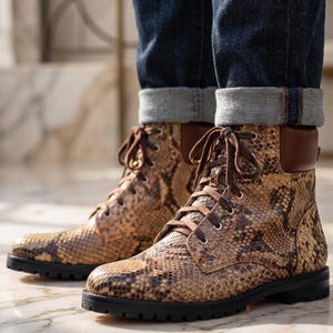 Puede incluir: Un par de botines con un patrón de piel de serpiente en tonos beige y marrón, cuellos de cuero marrón y cordones de color marrón oscuro. Las botas tienen ojales plateados y una suela de goma negra gruesa con tacos para mayor firmeza.