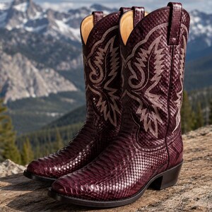 May include: A pair of deep burgundy cowboy boots featuring a textured snakeskin pattern throughout the shaft and vamp. The boots include light-colored decorative stitching in a wing-like design on the upper shaft and have a stacked heel.