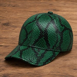 Puede incluir: Una gorra de béisbol con un patrón de textura verde y negra similar a la piel de serpiente. La gorra presenta una corona estructurada de seis paneles, una visera curva y un pequeño botón de tela a juego en la parte superior central.
