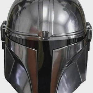 May include: A polished chrome-finish helmet featuring a smooth, rounded dome, a central vertical ridge, and a dark, T-shaped visor opening. The helmet has angular cheek plates and circular side ear caps, reflecting light across its metallic surface.