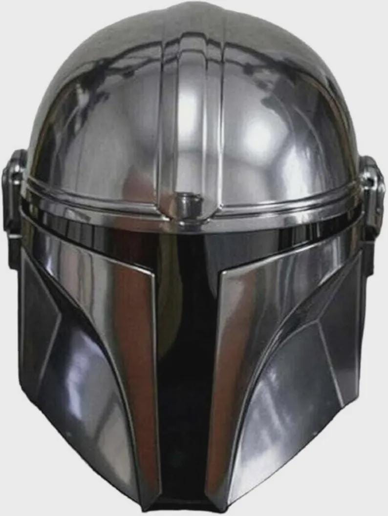 May include: A polished chrome-finish helmet featuring a smooth, rounded dome, a central vertical ridge, and a dark, T-shaped visor opening. The helmet has angular cheek plates and circular side ear caps, reflecting light across its metallic surface.