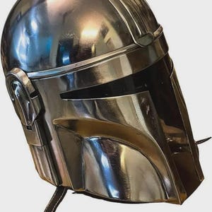 May include: A polished silver-colored metal helmet featuring a smooth, rounded dome, a T-shaped visor opening, and integrated side ear covers. The helmet has a reflective surface and includes a thin, dark strap hanging from the lower edge.