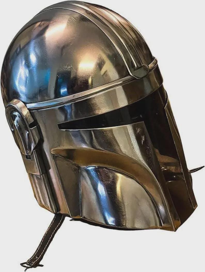 May include: A polished silver-colored metal helmet featuring a smooth, rounded dome, a T-shaped visor opening, and integrated side ear covers. The helmet has a reflective surface and includes a thin, dark strap hanging from the lower edge.