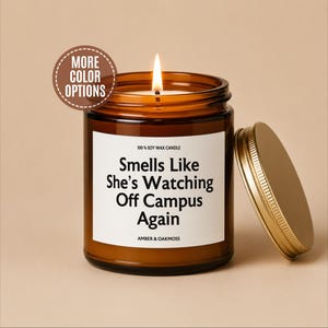 May include: A lit soy wax candle in an amber glass jar with a gold lid. The white label reads 100% SOY WAX CANDLE, Smells Like She’s Watching Off Campus Again, and AMBER & OAKMOSS. A brown circular badge in the top left corner says MORE COLOR OPTIONS.