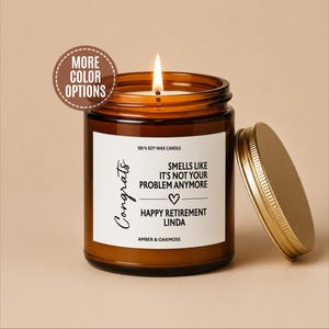May include: A lit soy wax candle in an amber glass jar with a gold lid. The white label reads: Congrats, Smells like it's not your problem anymore, Happy Retirement Linda, Amber & Oakmoss. A brown circular badge says More Color Options.