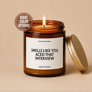 May include: A lit soy wax candle in an amber glass jar with a gold lid. The white label reads 100% SOY WAX CANDLE, SMELLS LIKE YOU ACED THAT INTERVIEW, and AMBER & OAKMOSS. A brown circular badge in the top left corner says MORE COLOR OPTIONS.