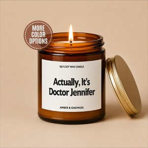 May include: A lit soy wax candle in an amber glass jar with a gold metal lid. The white label features the text 100% SOY WAX CANDLE, Actually, It's Doctor Jennifer, and AMBER & OAKMOSS. A circular badge in the top left corner reads MORE COLOR OPTIONS.