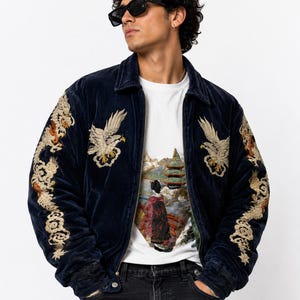 May include: A navy blue velvet bomber jacket featuring intricate gold and cream embroidery of eagles on the chest and dragons along the sleeves. The jacket is worn over a white t-shirt with a detailed graphic print of a landscape and a figure.