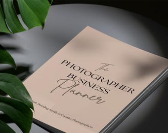 May include: A beige business planner with black text that reads The Photographer Business Planner and Portrait, Branding, Family & Creative Photographers. The cover has a matte finish and is positioned next to large green monstera leaves.