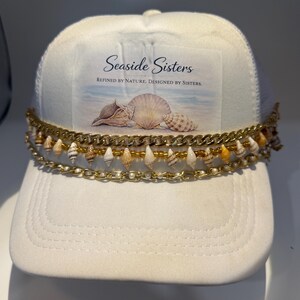 May include: A white baseball cap featuring a rectangular patch with the text Seaside Sisters, Refined by Nature, Designed by Sisters, and an image of seashells. The cap is adorned with two gold-tone chains and a strand of small seashells.