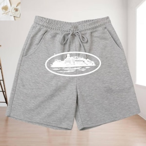 May include: Light grey sweat shorts featuring an elastic waistband with a drawstring closure and two side pockets. A white graphic print centred on the front depicts a cruise ship inside an oval frame, rendered in a simple line art style.