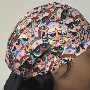 Peut inclure: Une charlotte de bloc opératoire avec un motif répétitif de personnages de dessins animés habillés en infirmiers avec des uniformes roses et bleus. Le tissu présente plusieurs visages aux grands yeux. Un bouton noir est fixé sur le côté.