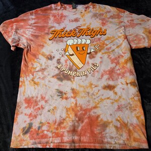 May include: A short-sleeve t-shirt with a tie-dye pattern in shades of orange, brown, and white. The center features a graphic of a cartoon pumpkin pie slice with a face, surrounded by the text Thick Thighs & Pumpkin Pies in brown lettering.