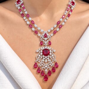 Puede incluir: Un collar de doble cadena con piedras rojas de corte ovalado y piedras blancas transparentes engastadas en metal plateado. La pieza central es un colgante ornamentado con una gran piedra roja de corte cojín, rodeada de piedras claras y gotas rojas.