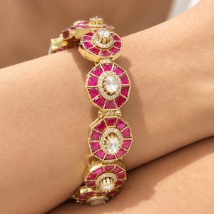 May include: A gold-toned bracelet featuring a series of connected octagonal links. Each link is set with vibrant pink stones arranged in a geometric pattern, surrounding a central oval white stone framed by small clear pavé crystals.