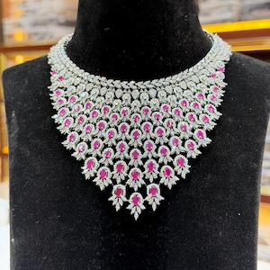 May include: A wide, ornate statement necklace featuring a dense arrangement of oval-cut vibrant pink stones and numerous small, clear, sparkling white stones set in a silver-toned metal frame, creating a layered, scalloped, and intricate design.