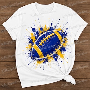 Puede incluir: Una camiseta blanca de manga corta con un gráfico central de un balón de fútbol americano. El balón es azul con rayas y cordones amarillos, rodeado por un diseño de salpicaduras de pintura azul y amarilla que irradia desde el centro.