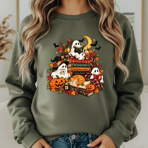 May include: A military green crewneck sweatshirt featuring a graphic print of three ghosts, a cat, pumpkins, and a stack of books with titles including Pumpkin Spice, Spooky Stories, Halloween Magic, and Good Books, surrounded by autumn leaves.