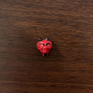 May include: A small, red, sculpted devil head charm featuring black painted horns, eyebrows, and a pointed goatee. The facial features include wide eyes with white accents, a prominent nose, and a subtle smile, all rendered in a smooth, molded material.