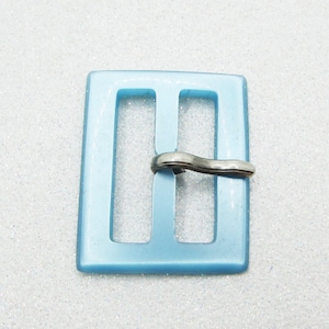 May include: A light blue, square buckle with a silver-colored clasp. The buckle has two vertical bars and a rectangular shape. The clasp is a curved, silver-toned metal piece.
