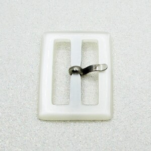 May include: A rectangular, off-white buckle with a silver-colored clasp. The buckle has two vertical bars and a central bar with a pivoting clasp. The background is a textured, white surface.
