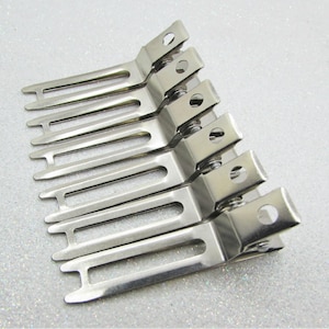 May include: A set of seven silver-colored metal hair clips. Each clip has a rectangular top with a circular hole and a series of parallel tines. The clips are arranged diagonally on a textured, light gray surface.