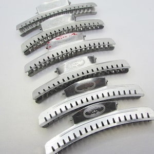 May include: A collection of vintage silver-toned hair barrettes. The barrettes have a curved design with a series of small holes along the edge. Some barrettes have an oval-shaped decorative element. The barrettes are arranged on a white surface.