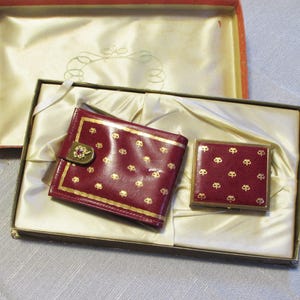 May include: A vintage burgundy wallet and compact set with gold accents and a decorative pattern, presented in a matching box. The wallet features a clasp with a jewel. The set is displayed on a satin-like fabric.