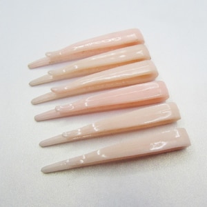 May include: Six pale pink hair barrettes are arranged on a white surface. Each barrette has a rectangular top and tapers to a pointed end. The barrettes are made of a smooth, glossy material.