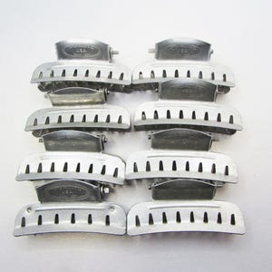 May include: Eight vintage silver-toned hair clips arranged in two columns. Each clip has a curved, slotted design with a hinged top and a small, stamped marking that reads "U.S.A.". The clips are likely from the mid-20th century.